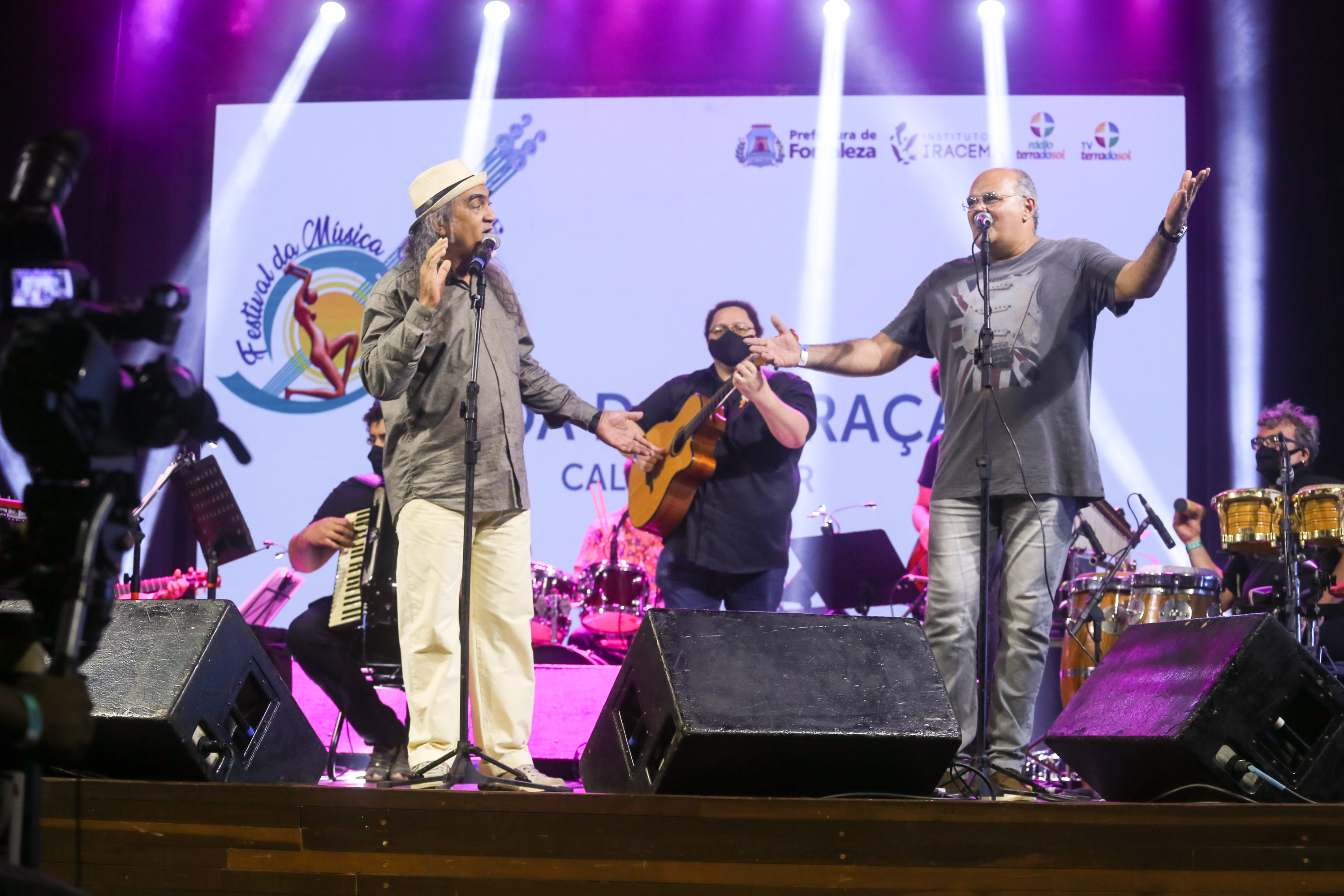 Dois homens cantando em pé e banda atrás tocando em cima de palco